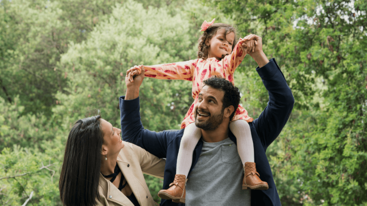 Father holding daughter on his shoulders while laughing and mother is looking up at daughter smiling